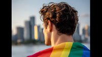 Young Caucasian man with curly dark hair wearing rainbow flag cape looking at city skyline during sunset, back view portrait celebrating pride and diversity.