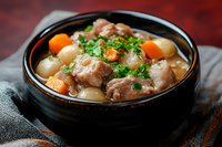 Hearty meat stew with tender pork chunks, pearl onions, carrots and fresh herbs served in traditional black ceramic bowl on rustic background.