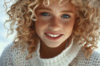 Young woman with natural freckles, bright blue eyes and curly blonde hair wearing white knit sweater smiles genuinely at camera, radiating warmth and authenticity.