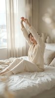 Young Asian woman in cream pajamas stretching arms above head while sitting on bed in bright bedroom with natural morning light streaming through curtains.