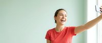 Happy young Caucasian woman in red t-shirt taking selfie with smartphone, smiling joyfully in bright modern interior with mint green wall background.