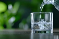 Pouring pure water from plastic bottle into glass tumbler with splashing liquid against blurred green nature background, refreshing drink concept.
