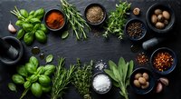 Fresh herbs and spices arranged on dark slate background with basil, rosemary, bay leaves, peppercorns, sea salt, paprika and garlic for cooking.