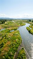 Aerial view of winding river through lush green valley with forested banks, agricultural fields, and distant mountains under clear sky. Perfect for environmental and landscape projects.