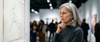 Mature woman with gray hair contemplating artwork in modern gallery space with visitors in background.