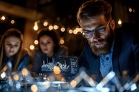 Focused businessman with beard and glasses analyzing financial data charts on digital tablet during evening team meeting with warm ambient lighting.