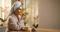 Young Asian woman in bathrobe and towel applying golden face mask in bright bathroom with orchids for skincare routine and spa wellness at home.