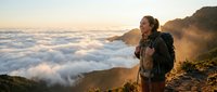 Woman hiker with backpack standing on mountain peak above clouds at sunrise, enjoying scenic landscape view and outdoor adventure.
