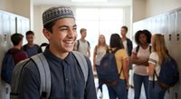 Happy young Middle Eastern male student with traditional cap smiling in busy school hallway with diverse classmates carrying backpacks during break time.