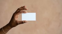 Close-up of a hand with medium-dark skin tone holding a blank white card against a neutral beige background, suitable for business card mockups and branding presentations.