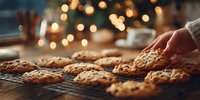 Freshly baked chocolate chip cookies cooling on wire rack with hand reaching for one, Christmas tree lights bokeh in background.