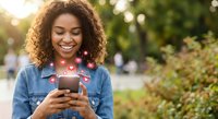 Happy young African American woman using smartphone with heart emojis floating around screen, smiling while texting outdoors in sunny park setting.