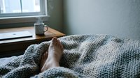 Cozy morning scene with bare feet under soft knitted blanket in bed, coffee mug on windowsill creating peaceful hygge atmosphere for relaxation and comfort.