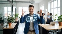 Young man with light skin tone and dark hair wearing casual business attire raising both hands in celebration within a modern office environment with colleagues and plants, expressing success and achievement.