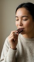 Young Asian woman enjoying dark chocolate piece with closed eyes in cozy cream sweater, expressing pleasure and mindful eating moment for wellness content.