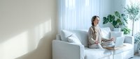 Young professional woman working remotely on laptop in bright modern living room with plants and natural light streaming through windows.