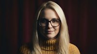 Young blonde Caucasian woman wearing glasses and yellow turtleneck sweater smiling confidently in warm indoor lighting for professional headshot photography.