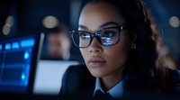 Young African American businesswoman in glasses working late at office, focused on computer screen with blue technology interface reflection.