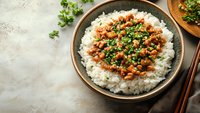 Spicy minced chicken with fresh herbs served over steamed white rice in ceramic bowl, garnished with chopped cilantro and green onions on light concrete background.