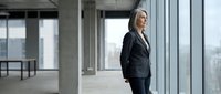 Mature woman in business attire standing by large windows in modern office space, looking out at cityscape. Corporate professional environment.
