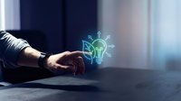 Businessman hand touching glowing lightbulb hologram on desk representing innovation creativity and bright ideas in modern office environment.