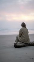 Woman with long gray hair sitting on driftwood log at beach during twilight, wearing neutral cardigan, gazing toward calm ocean horizon at sunset or sunrise.