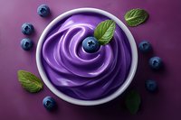 Purple whipped cream dessert topped with fresh blueberries and mint leaves in white ceramic bowl on dark background, overhead view of sweet treat.