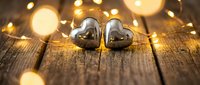 Two silver metallic heart shapes on rustic wooden surface with warm golden bokeh lights creating romantic Valentine's Day atmosphere for love concepts.