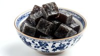 Black grass jelly dessert cubes in traditional Asian blue and white porcelain bowl with floral pattern on white background, cooling summer treat.