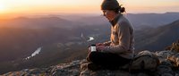 Male hiker in casual outdoor gear sitting on mountain peak at sunset, reviewing map while planning route through valley landscape below.