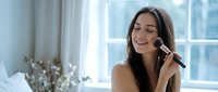Young brunette woman applying makeup with brush in bright bathroom mirror, morning beauty routine skincare cosmetics preparation lifestyle.