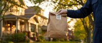 Delivery person holding brown paper bag with food order outside residential home during golden hour sunset for takeout and meal delivery service.