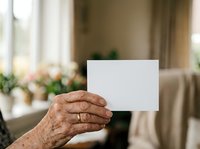Senior woman holding blank white card in living room with natural window light and blurred home interior background for greeting, announcement, or message display.