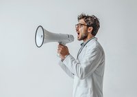 Young Caucasian male doctor in white coat and glasses shouting through megaphone against light background, expressing urgent medical announcement or protest.
