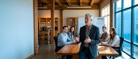 Senior male professional with gray hair and beard, wearing business casual attire, leading a meeting in a modern office with wooden beams and large windows. Diverse team of colleagues seated at table listening attentively. Corporate training, leadership and business communication concept.