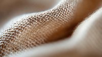 Natural linen fabric with visible weave texture in beige and cream tones creates soft waves and folds, macro close-up view showing detailed textile structure.