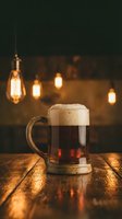 Glass mug of dark beer with foam on wooden table in cozy pub with warm Edison bulb lighting creating atmospheric ambiance for relaxation and socializing.