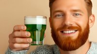 Young caucasian man with red beard and bright smile holding glass of green beer, celebrating St Patrick's Day holiday in casual grey sweater.