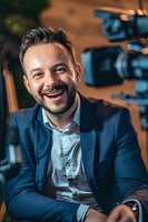 Caucasian male professional in navy blue suit laughing genuinely during video production. Professional cameras in background create cinematic atmosphere.