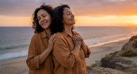 Two happy women embracing on beach at sunset wearing matching brown shirts with ocean waves and golden sky creating romantic peaceful moment for friendship.