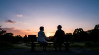 Two people sitting on a bench at sunset, silhouetted against a golden sky with trees and rural landscape, suitable for themes of friendship, relaxation, and outdoor leisure.
