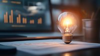 Glowing light bulb on business documents with financial charts in background, symbolizing innovative business solutions and creative ideas in office setting.