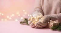 Woman in cozy pink sweater holding glowing snow globe with golden lights and gift boxes on soft pink background with bokeh lights creating magical holiday atmosphere.