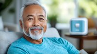 Smiling senior Asian man with white beard wearing turquoise t-shirt relaxing on gray couch at home, expressing joy and contentment in comfortable living room setting.