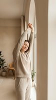 Young woman with medium skin tone stretching arms overhead in bright hallway wearing beige knit sweater and linen pants, morning wellness and relaxation concept for lifestyle and health content.