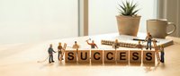 Miniature figures assembling wooden blocks spelling success on desk with plant and coffee cup, representing teamwork, achievement and business growth.