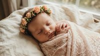 Peaceful newborn baby girl sleeping wrapped in soft beige blanket wearing delicate floral crown headband for portrait photography sessions.