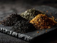 Assorted loose tea leaves in piles on dark stone slate, showcasing black, green, and golden varieties for gourmet beverage preparation.