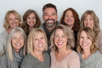 Diverse group of smiling adults with various hairstyles wearing neutral-colored sweaters and tops posing together against light background for portrait photography.