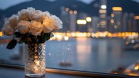 Romantic cream roses in sparkling glass vase against evening cityscape with blurred bokeh lights, creating intimate atmosphere on windowsill at dusk.